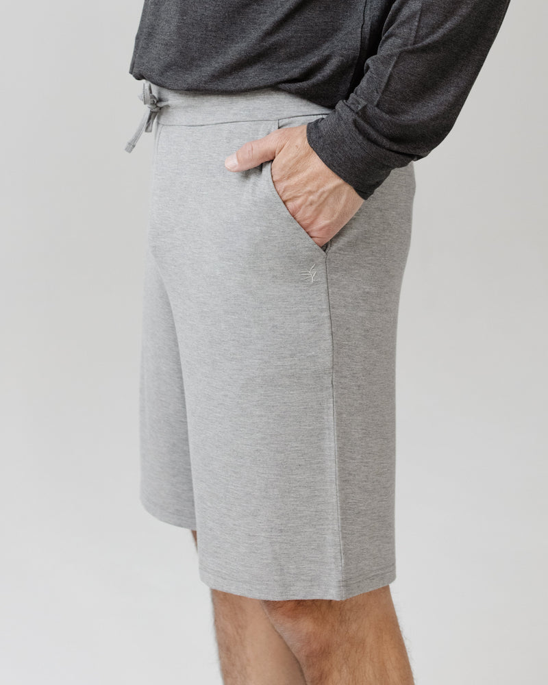 A man wearing the Cozy Earth Men's Bamboo Stretch-Knit Short Sleeve Pajama Set in light gray stands sideways against a plain light background, placing a hand in his pocket.