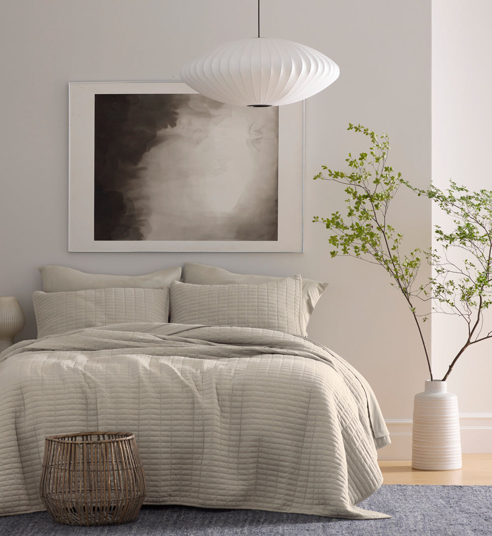 A bed featuring Cozy Earth Bamboo Jersey bedding in a bedroom with white walls, a tree in a planter, a blue rug, and a large abstract painting.