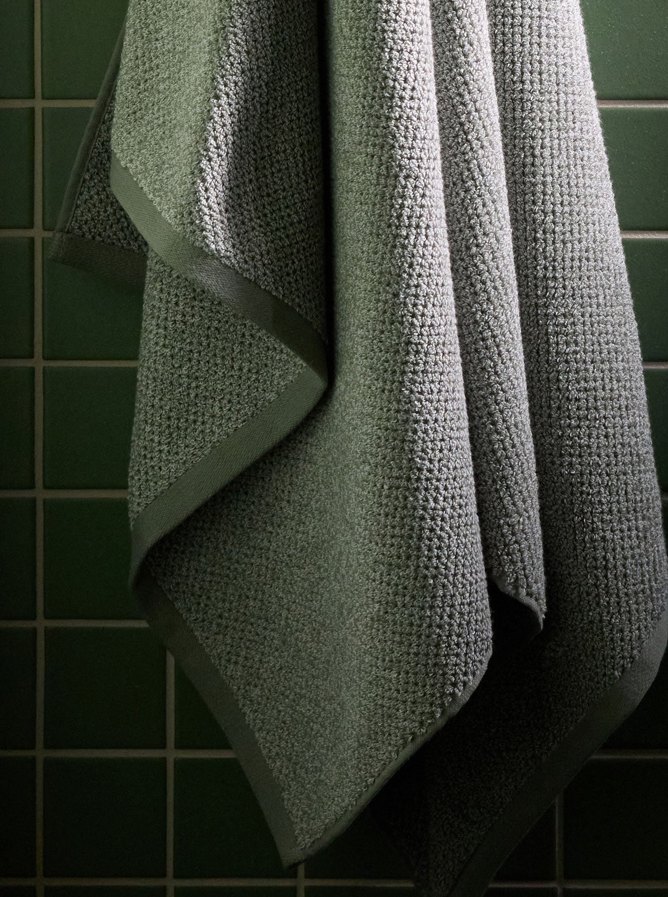 A Nantucket Bath Sheet in Heathered Sage is pictured hanging against a green tiled wall. 