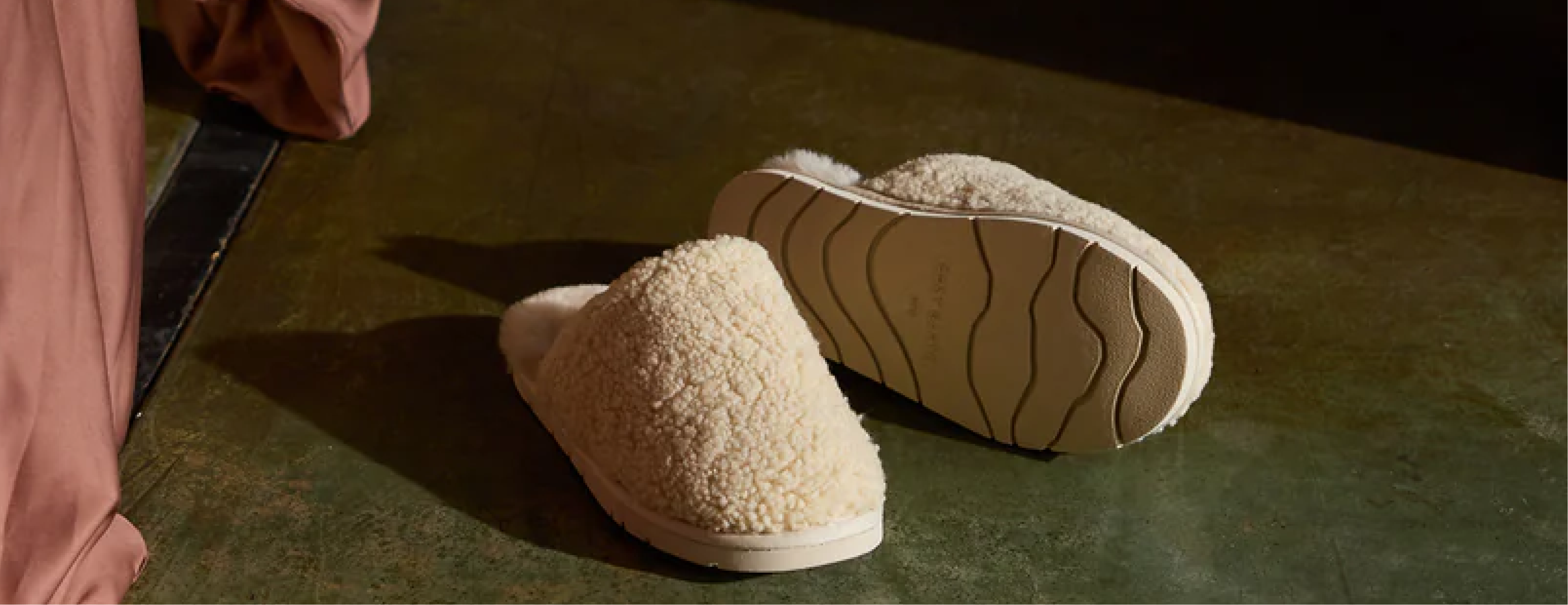 A pair of cozy, off-white textured slippers rests on a polished green floor, casting soft shadows. A pink curtain drapes nearby, partially visible at the edge of the frame.