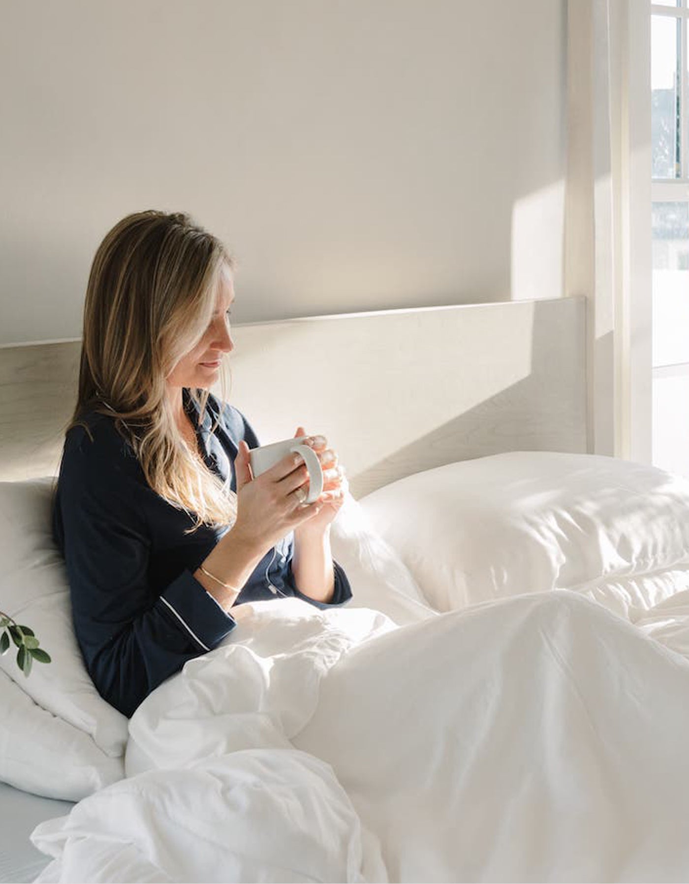 A woman with light hair sits up in bed, smiling in dark pajamas while holding a white mug. She’s surrounded by plush white Cozy Earth Silk Comforter bedding, bathed in sunlight from a window nearby.