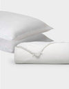 Two white pillows from Cozy Earth's Silk Insert Bundle are stacked diagonally behind a rolled-up white duvet with a visible tie closure, all set against a plain light gray background.|Filling:Standard