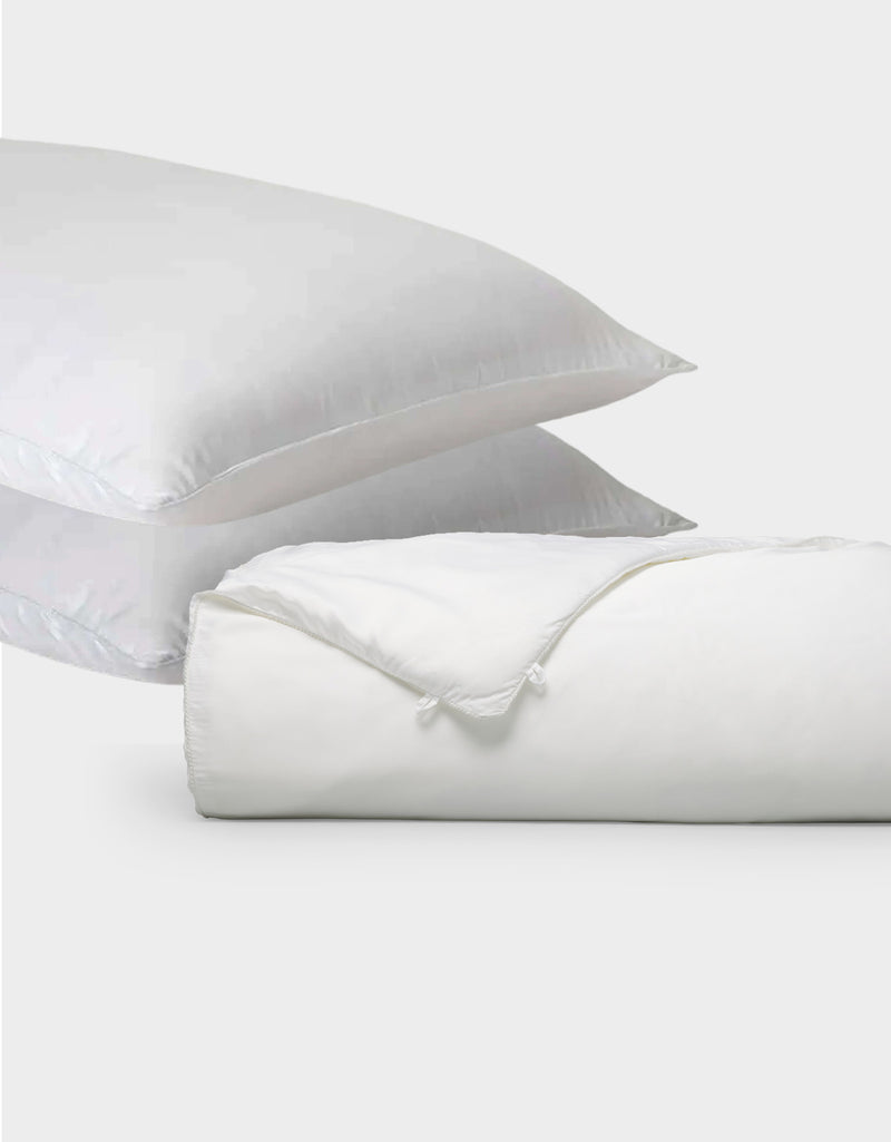 Two white pillows from Cozy Earth's Silk Insert Bundle are stacked diagonally behind a rolled-up white duvet with a visible tie closure, all set against a plain light gray background.