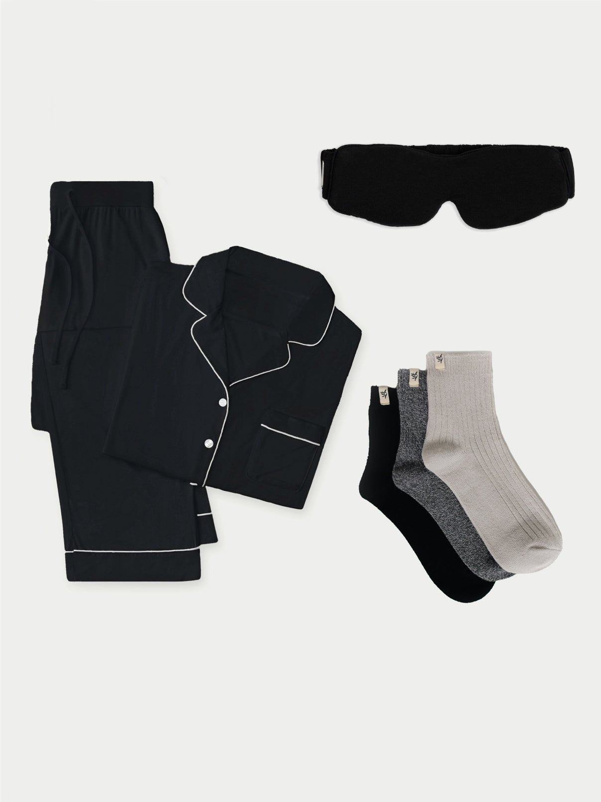 The Women's Bedtime Bundle by Cozy Earth is laid out flat. The bundle, in black, includes a long-sleeved shirt and pants with white piping. Next to them is a black sleep mask. To the right, there are three pairs of socks: one beige, one black, and one gray. 