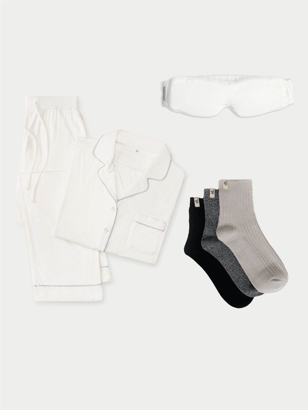 Introducing the Women's Bedtime Bundle by Cozy Earth: a neatly arranged set featuring white pajamas with a button-up top and pants, a matching white eye mask, and three pairs of socks in black, gray, and light gray, all beautifully displayed on a light background. 