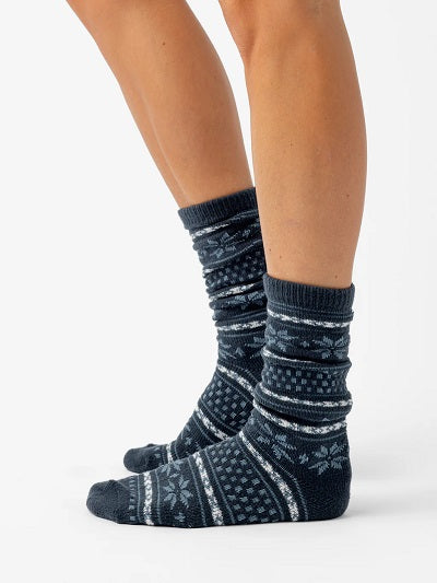 A person wearing Cozy Earth's The Snowflake Plush Lounge Sock - Last Chance in dark blue with white and light blue Nordic patterns stands on a white background, showing their lower legs and feet in a side view.