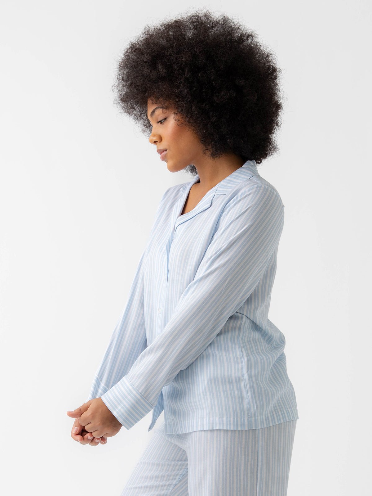 A person with curly hair, seen in profile against a plain background, wears the Women's Soft Woven Long Sleeve Pajama Set by Cozy Earth in light blue and white stripes. They are standing relaxed with gently clasped hands.