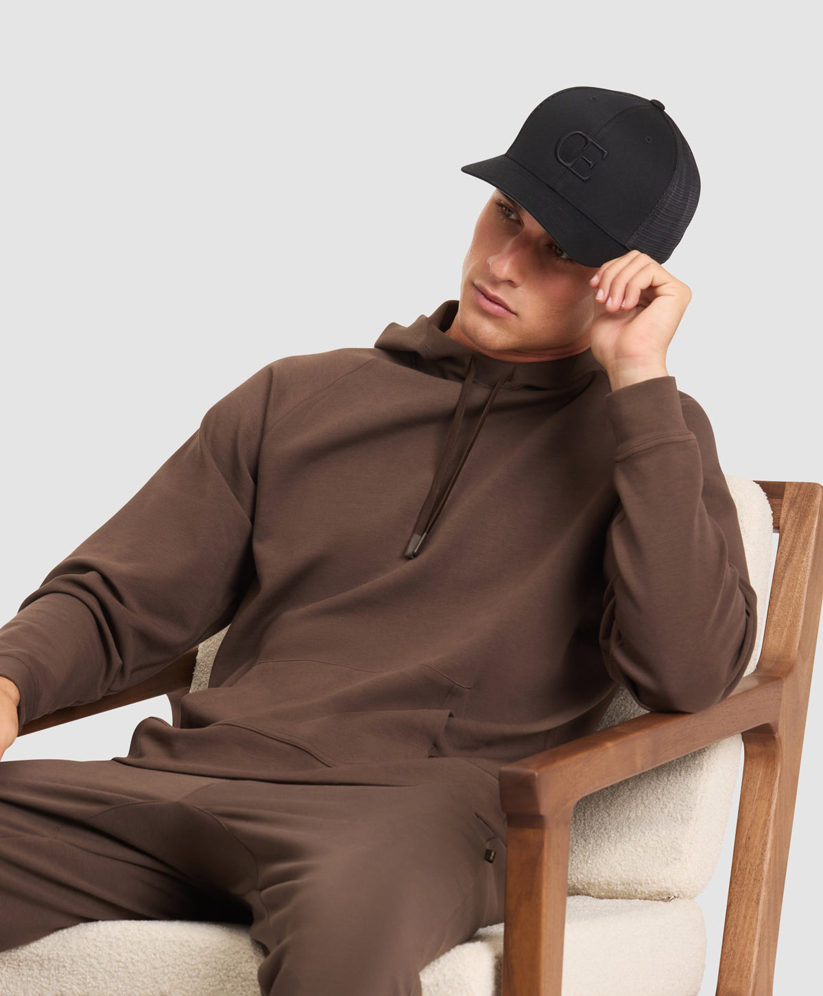 A man wearing a brown Cozy Earth StretchTech Hoodie and Joggers and black cap sitting on a chair against a light gray background