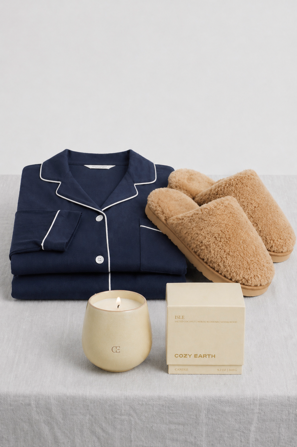 The Cozy Earth Unwind Bundle, featuring a navy pajama set with white piping, tan fuzzy slippers, and a tan candle in a glass container with matching box, is displayed on a light gray fabric surface.