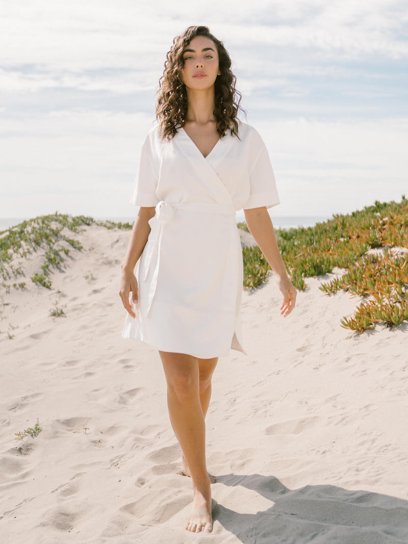 A person with curly hair walks barefoot on a sandy beach path, adorned in the Women's Cozy Earth Coastal Comfort Wrap Dress by Cozy Earth. The sky is partly cloudy, and the path is flanked by lush green shrubs and plants.