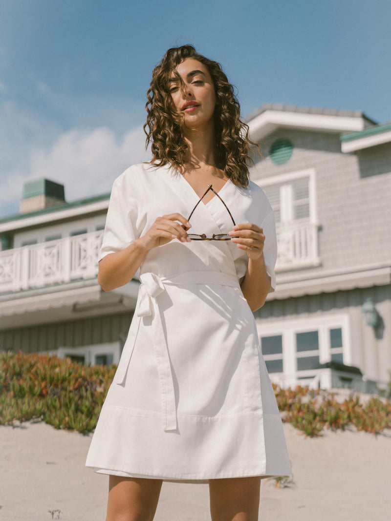 A woman with curly hair is standing in front of a house. She is wearing a White Women's Cozy Earth Coastal Comfort Wrap Dress by Cozy Earth and holding a pair of sunglasses in her hands. The background features a two-story house with a porch and railing, set against a partly cloudy sky.