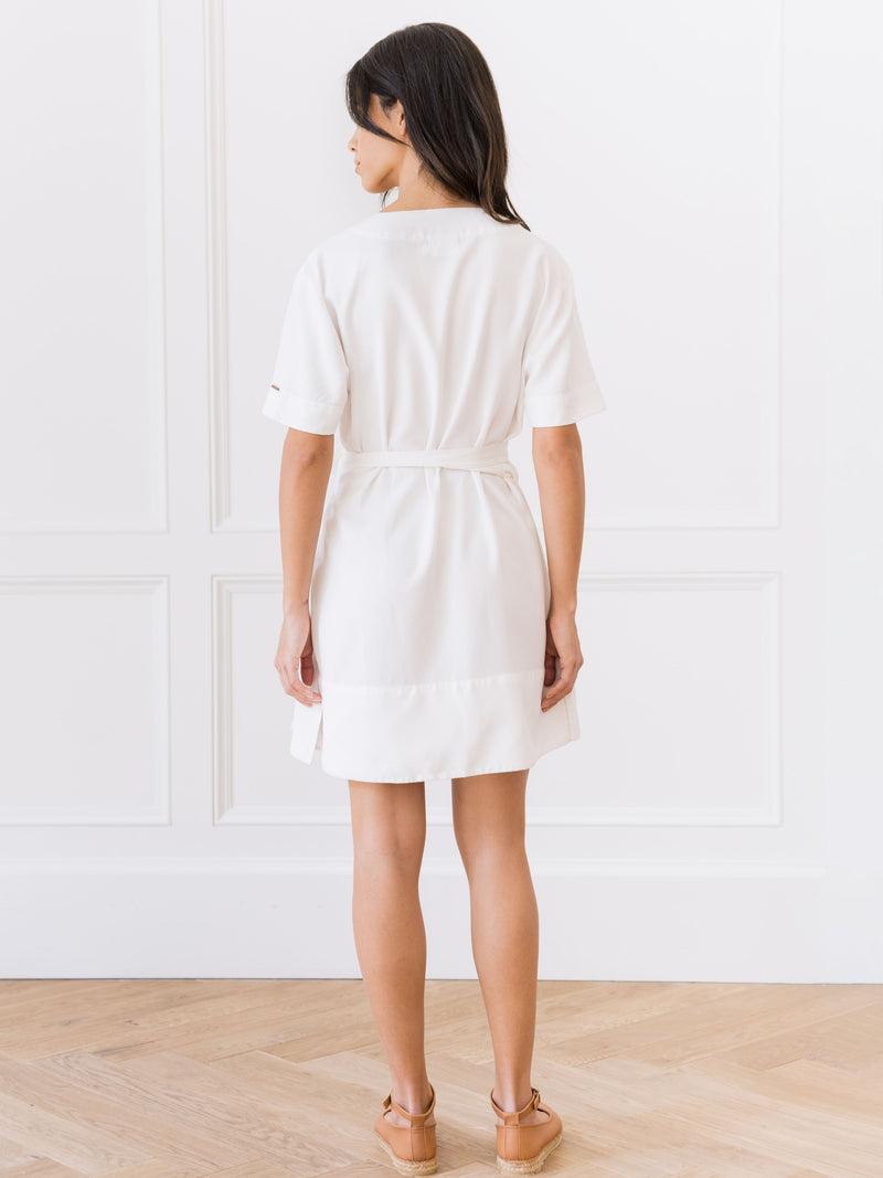 A person with long dark hair stands facing away, wearing the Women's Cozy Earth Coastal Comfort Wrap Dress from Cozy Earth with brown shoes. They are in a room with white paneled walls and a hardwood floor.