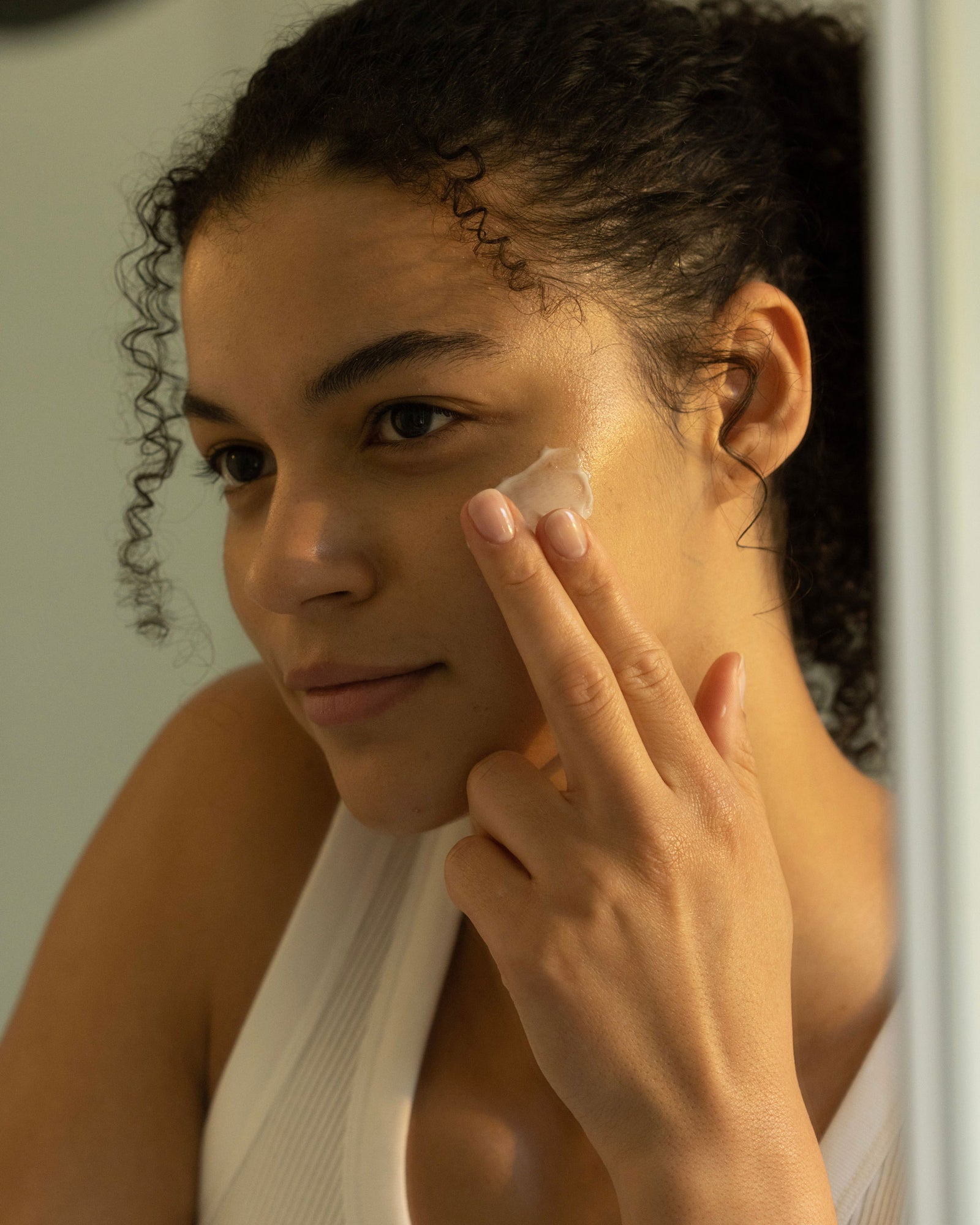 While looking in a mirror, a person with curly hair wearing a white top applies Cozy Earth's Mini Moisture Reset Trio to their cheek with their fingers under warm, natural lighting.