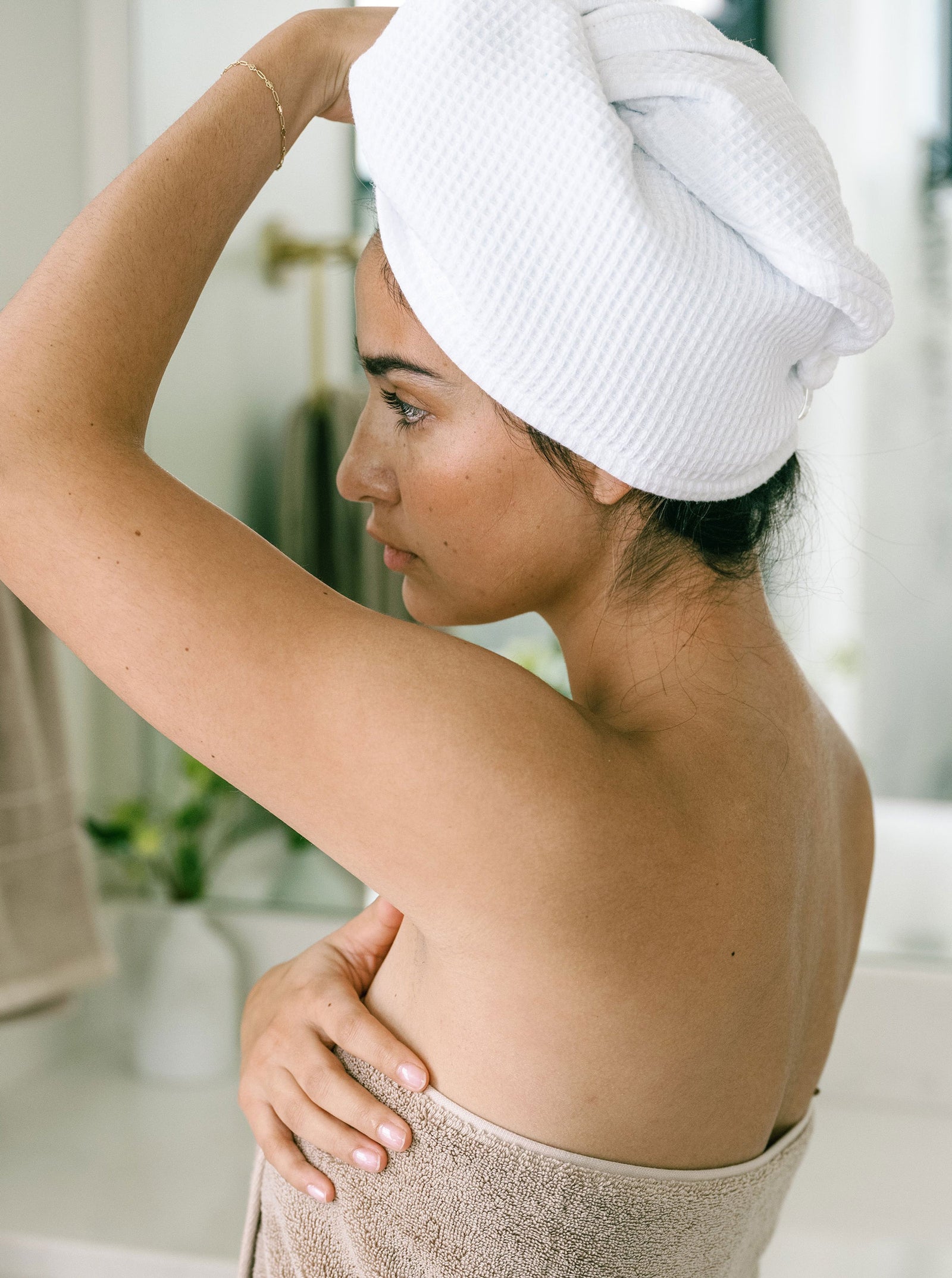 A woman in a bathroom setting, with the Cozy Earth Waffle Hair Towel wrapped around her head and a beige towel on her body, looks into a mirror, gently touching her hair. The softly blurred background hints at plants and bathroom fixtures.