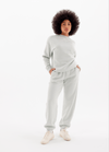 A woman with curly hair stands confidently in Cozy Earth's Women's CityScape Crewneck & Sweatpant Set in light gray, hands in pockets and paired with white sneakers, against a plain white background.|Color:Arctic