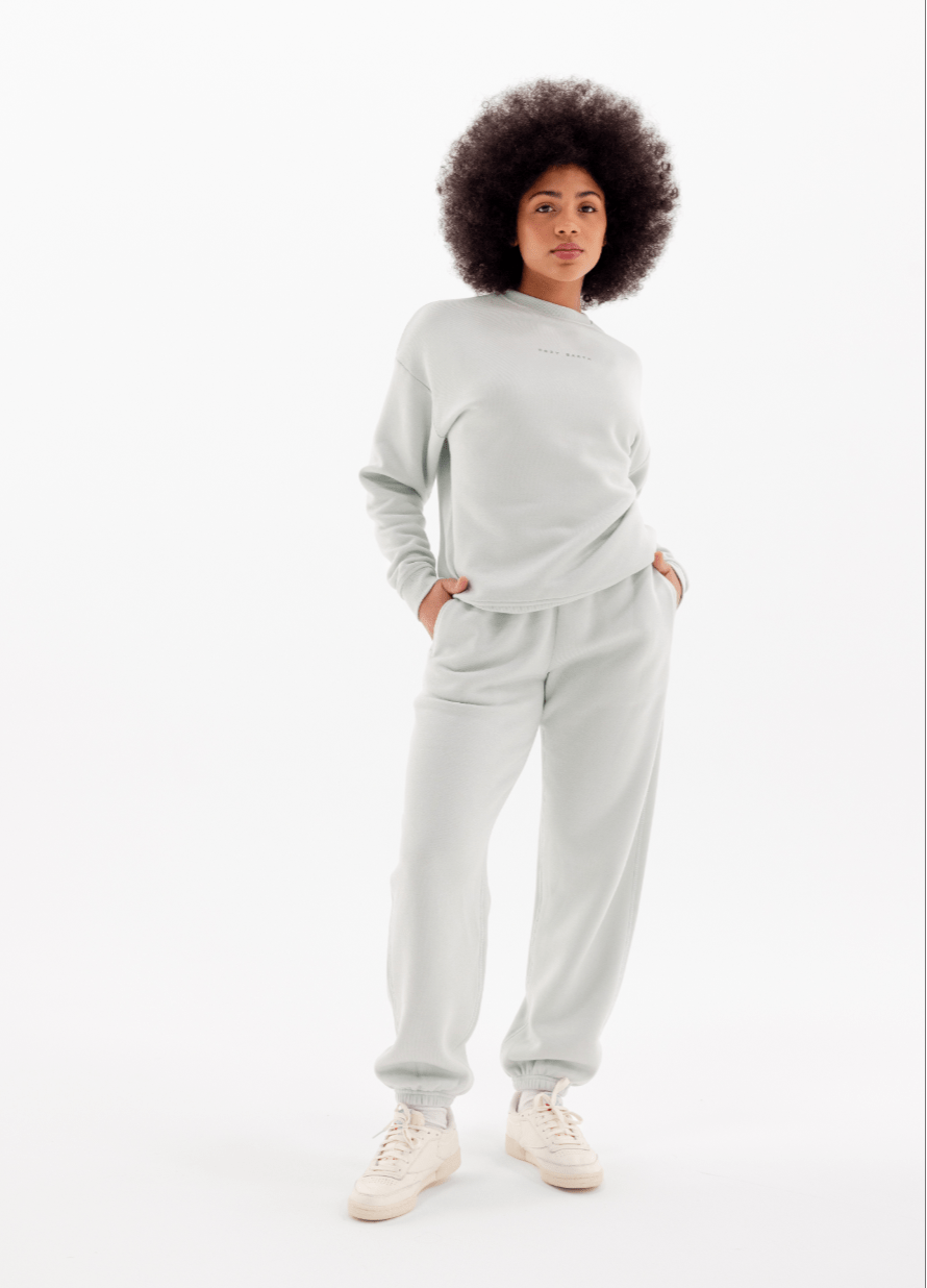 A woman with curly hair stands confidently in Cozy Earth's Women's CityScape Crewneck & Sweatpant Set in light gray, hands in pockets and paired with white sneakers, against a plain white background.|Color:Arctic