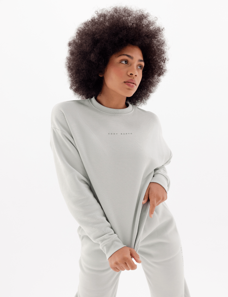 A woman with curly hair models the Cozy Earth Women's CityScape Crewneck & Sweatpant Set in light gray, standing against a white background and looking to the side while holding the hem of her sweatshirt.