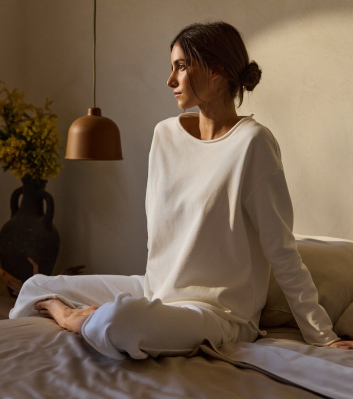 A woman sits on a bed in Cozy Earth Chelsea Set in white. A vase of flowers and a pendant lamp are in the background