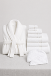The Cozy Earth Wrap Up Bundle, featuring a white bathrobe, assorted white towels, and a spa headband, is displayed on a light gray surface with a simple background.