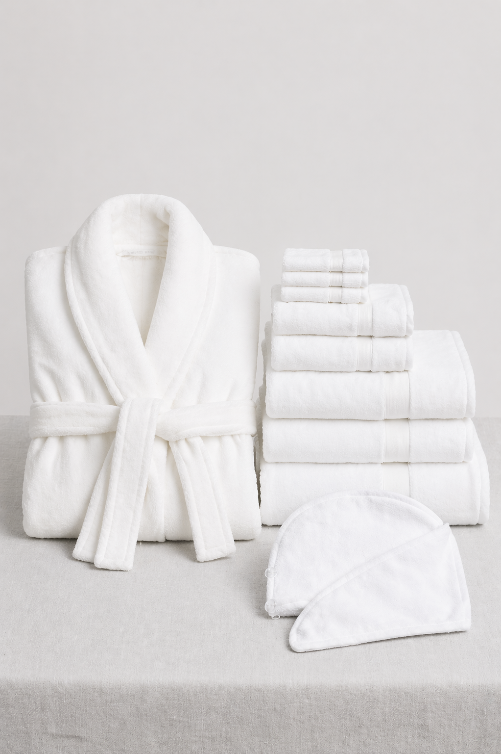 The Cozy Earth Wrap Up Bundle, featuring a white bathrobe, assorted white towels, and a spa headband, is displayed on a light gray surface with a simple background.