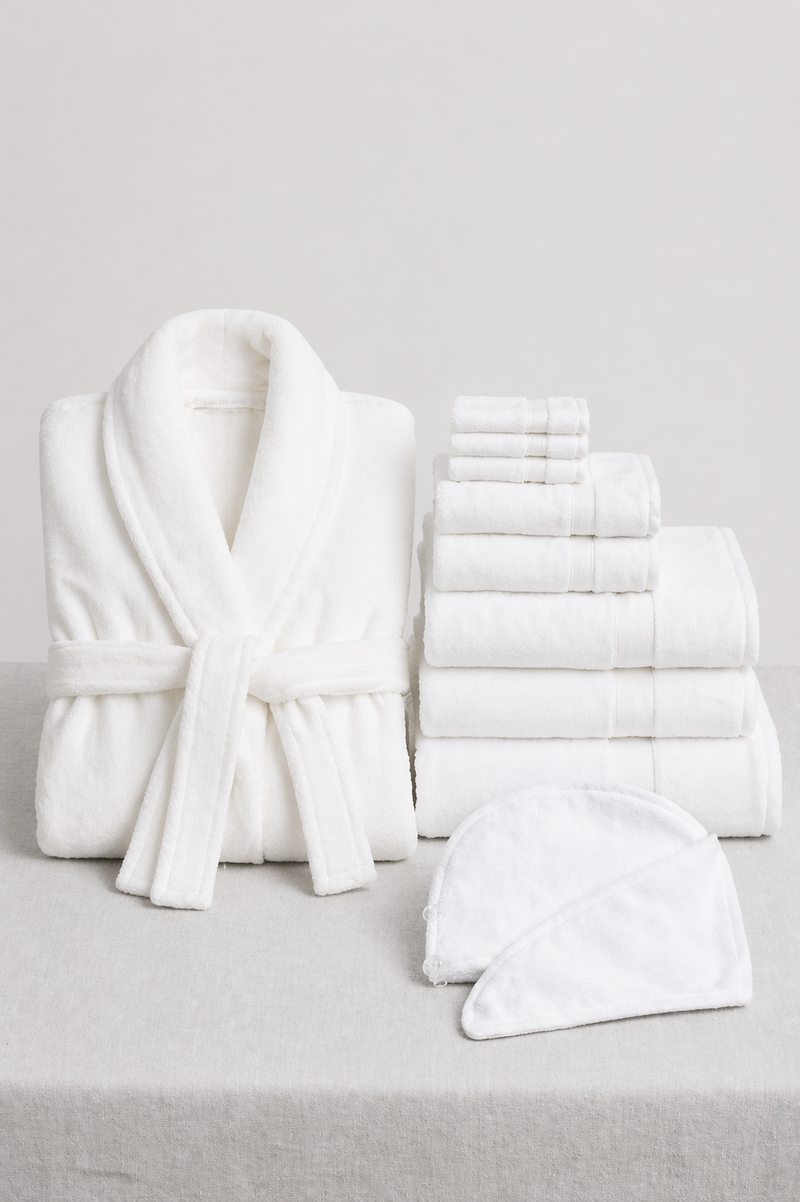 The Cozy Earth Wrap Up Bundle, featuring a white bathrobe, assorted white towels, and a spa headband, is displayed on a light gray surface with a simple background.