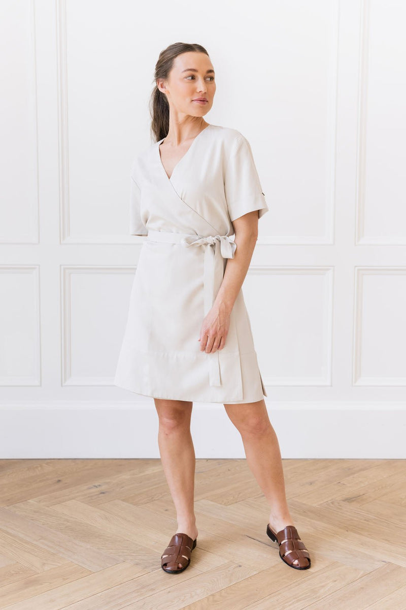A woman stands in a white room with wooden flooring. She is wearing the Cozy Earth Women's Coastal Comfort Wrap Dress in Sand, paired with brown sandals. She gazes off to the side with her hands relaxed at her sides.