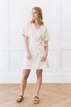 A woman with long, light brown hair stands in a room with white paneled walls and wooden flooring. She is wearing the Women's Cozy Earth Coastal Comfort Wrap Dress in Sunkissed Gingham, tied at the waist, paired with brown sandals. She gazes to her left with a neutral expression. |Color:Sunkissed Gingham