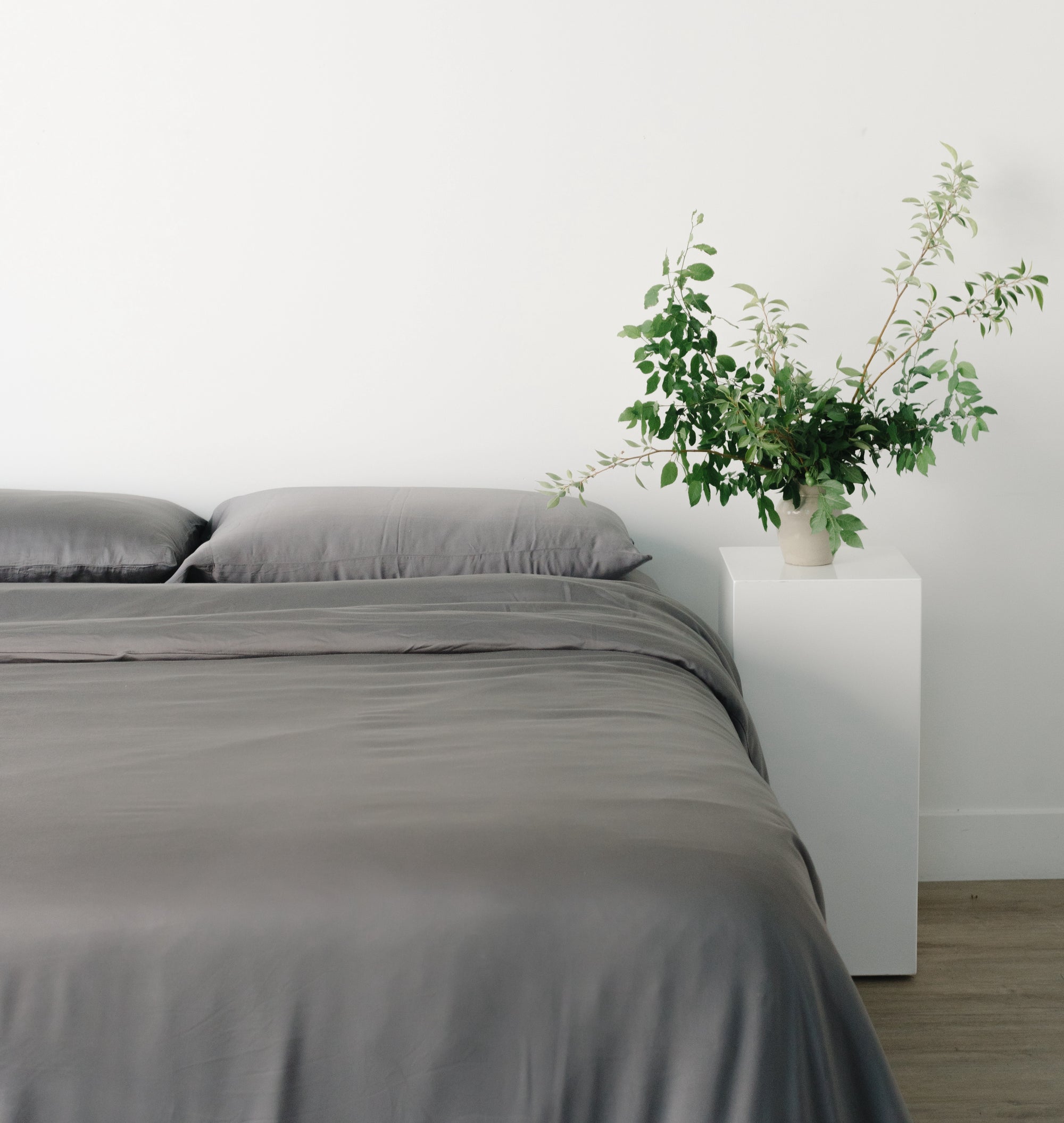 Bed made with charcoal bedding with a plant on the nightstand next to it 