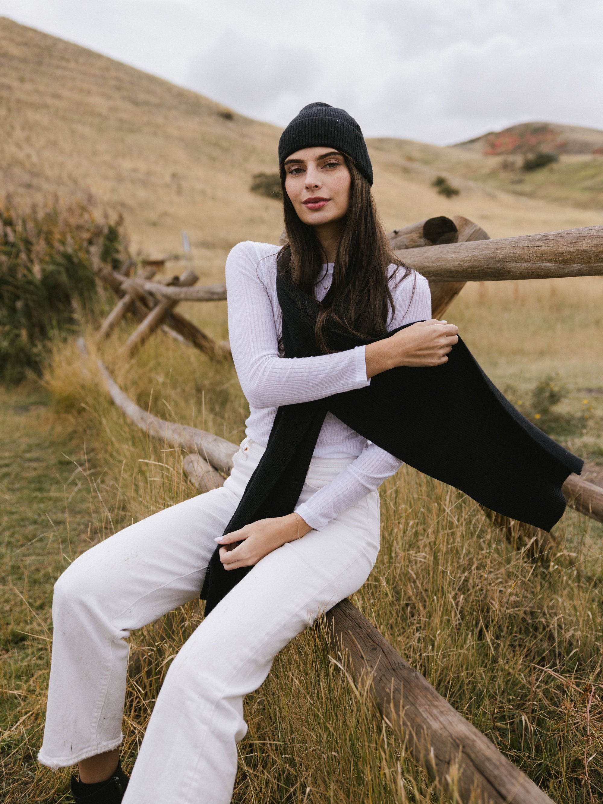 Woman wearing a black knit beanie and scarf sits on a wooden fence in a field 