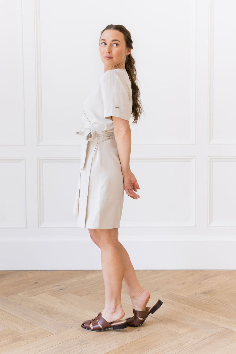 A woman wearing the Women's Cozy Earth Coastal Comfort Wrap Dress from Cozy Earth and brown sandals stands with her body turned slightly to the side. She has long brown hair tied back and holds her hands behind her. The background features a white paneled wall and light wooden flooring.
