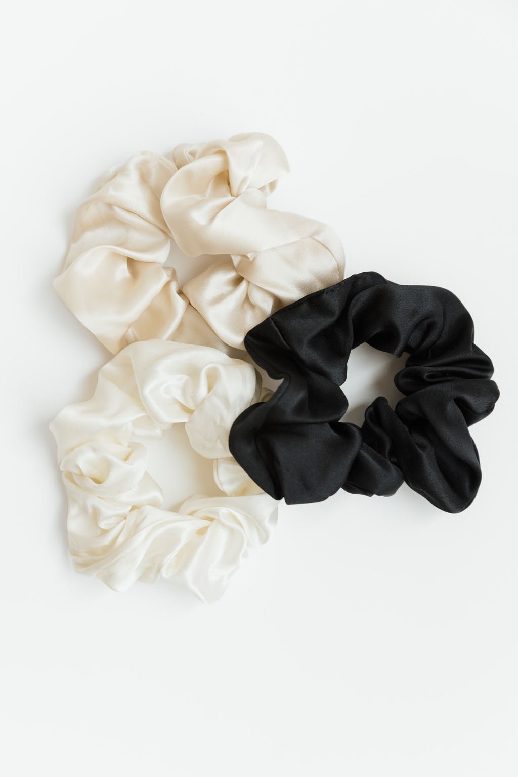 Three Silk Scrunchies from Cozy Earth are arranged in a triangle on a white background. Two scrunchies are cream-colored, and one is black. The soft, shiny fabric gives them a luxurious appearance.