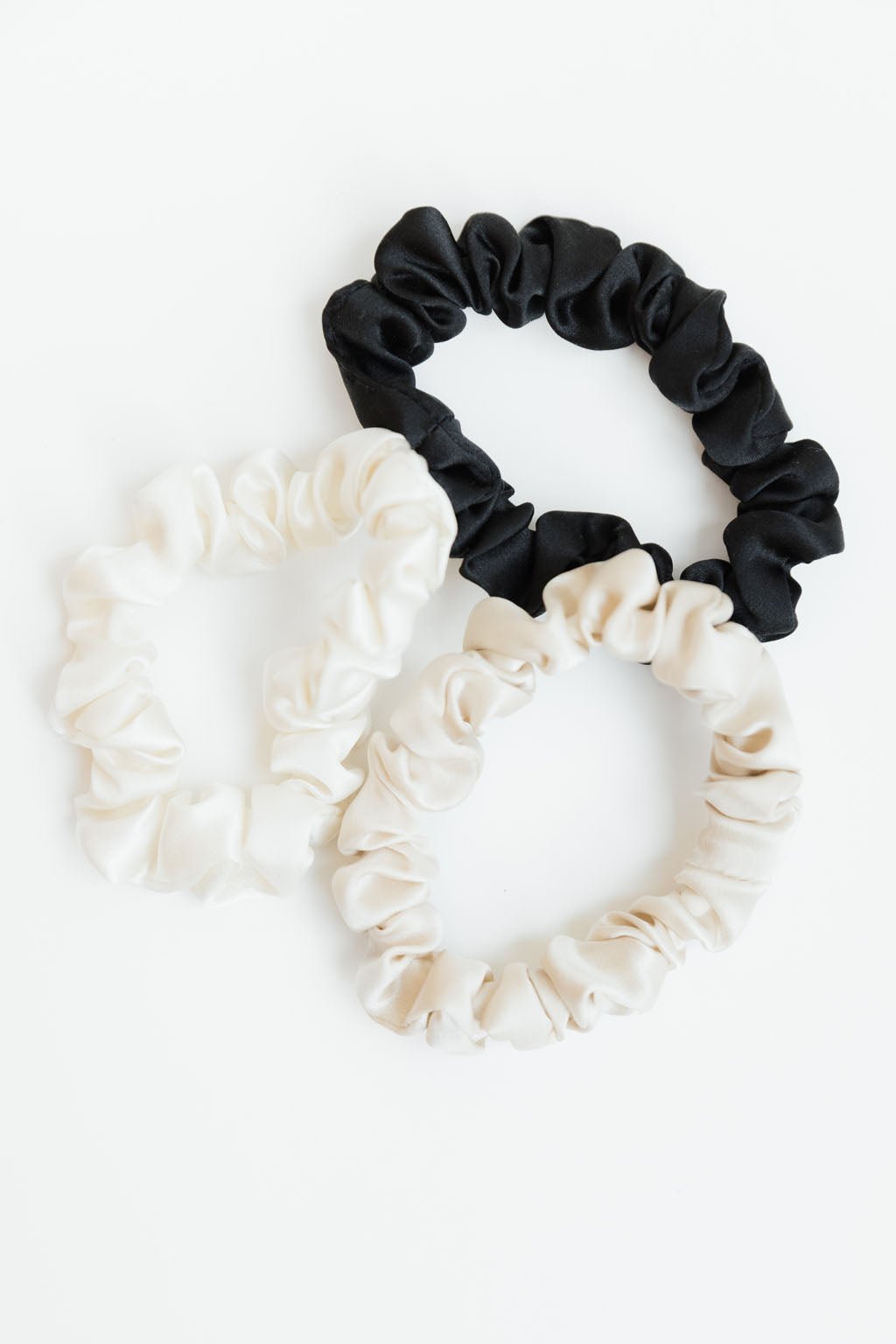 Three Silk Skinny Scrunchies from Cozy Earth are placed on a white background. Two of the scrunchies are cream-colored, and one is black. All of them have a ruffled texture. The scrunchies are arranged in such a way that they overlap slightly, forming a loose triangle.