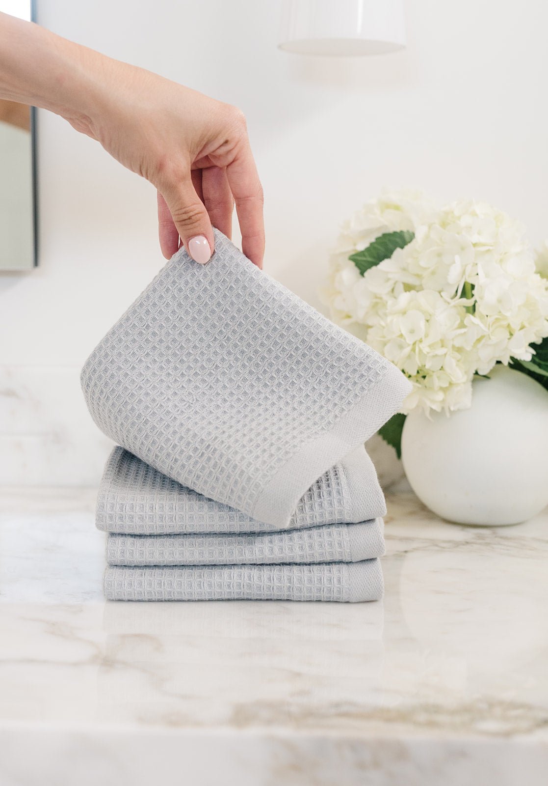 Harbor Mist Waffle Wash Cloths neatly folded. The photo was taken with a hand pulling one of the waffle cloths from the set. The cloths rest on a white marble sink 