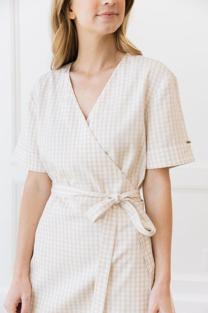 A woman in a beige and white checkered Cozy Earth Coastal Comfort Wrap Dress from Cozy Earth stands against a white background. The dress features short sleeves and a waist tie. Only the upper part of her body is visible, with her face partially out of the frame.