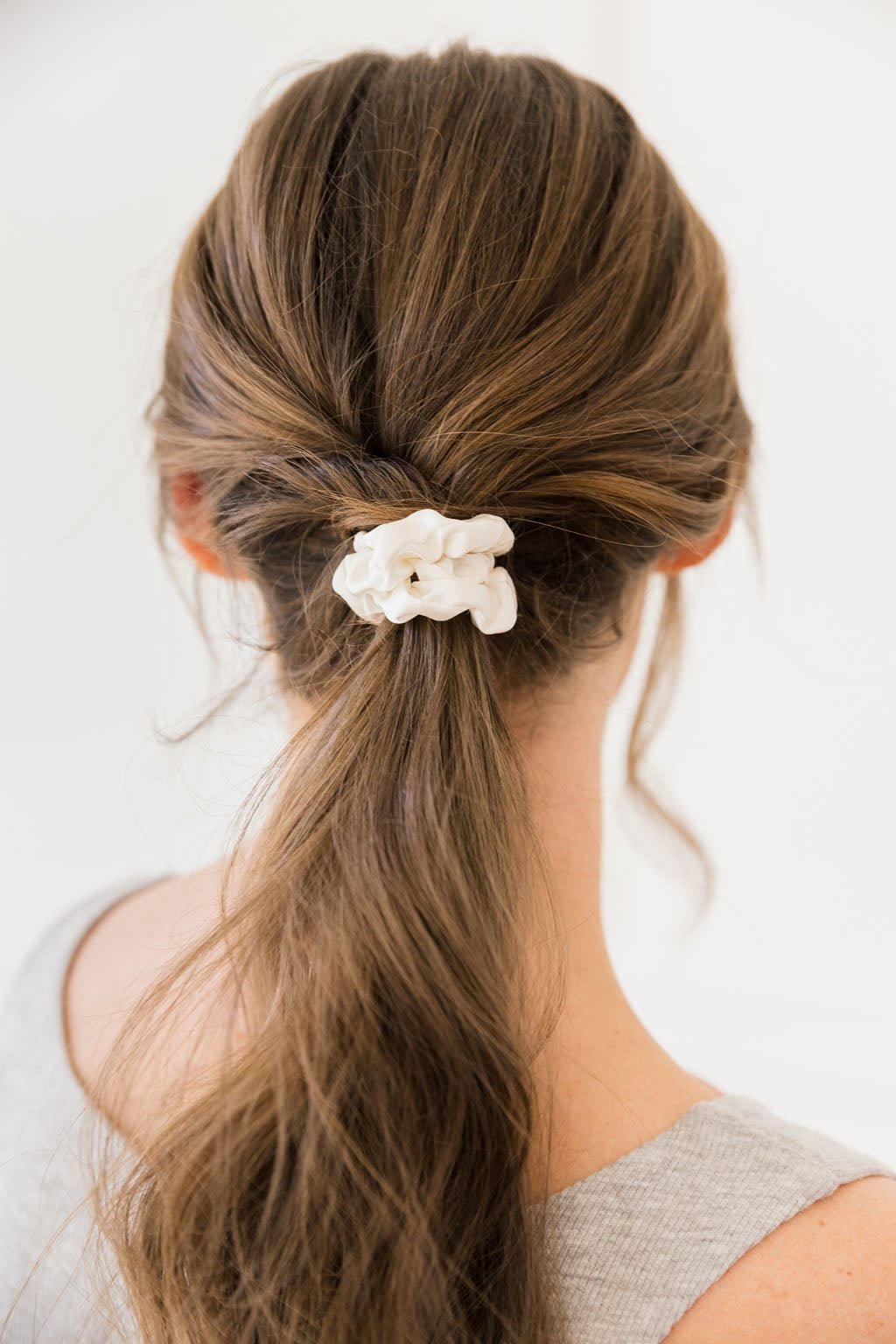 A person with long brown hair styled into a low, loose ponytail secured by a Cozy Earth Silk Skinny Scrunchie. The individual is wearing a light gray sleeveless top and is photographed from the back against a blurred, light background.