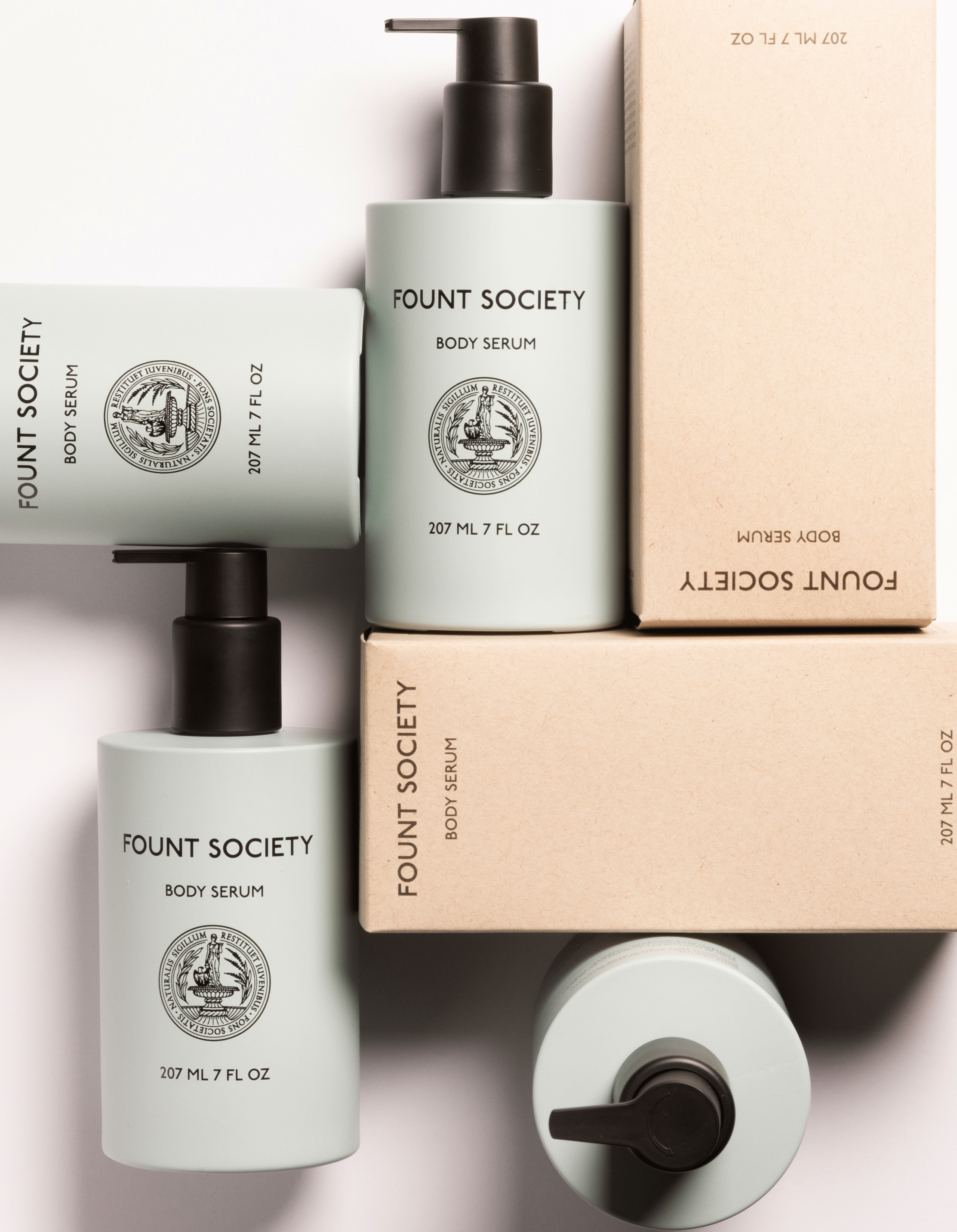 Arrangement of Cozy Earth Body Serum bottles, each 207 ml/7 fl. oz., with minimalist labels featuring a circular emblem. Some bottles lie horizontally while others stand upright. Two cardboard boxes are also included. The scene is bright and clean.