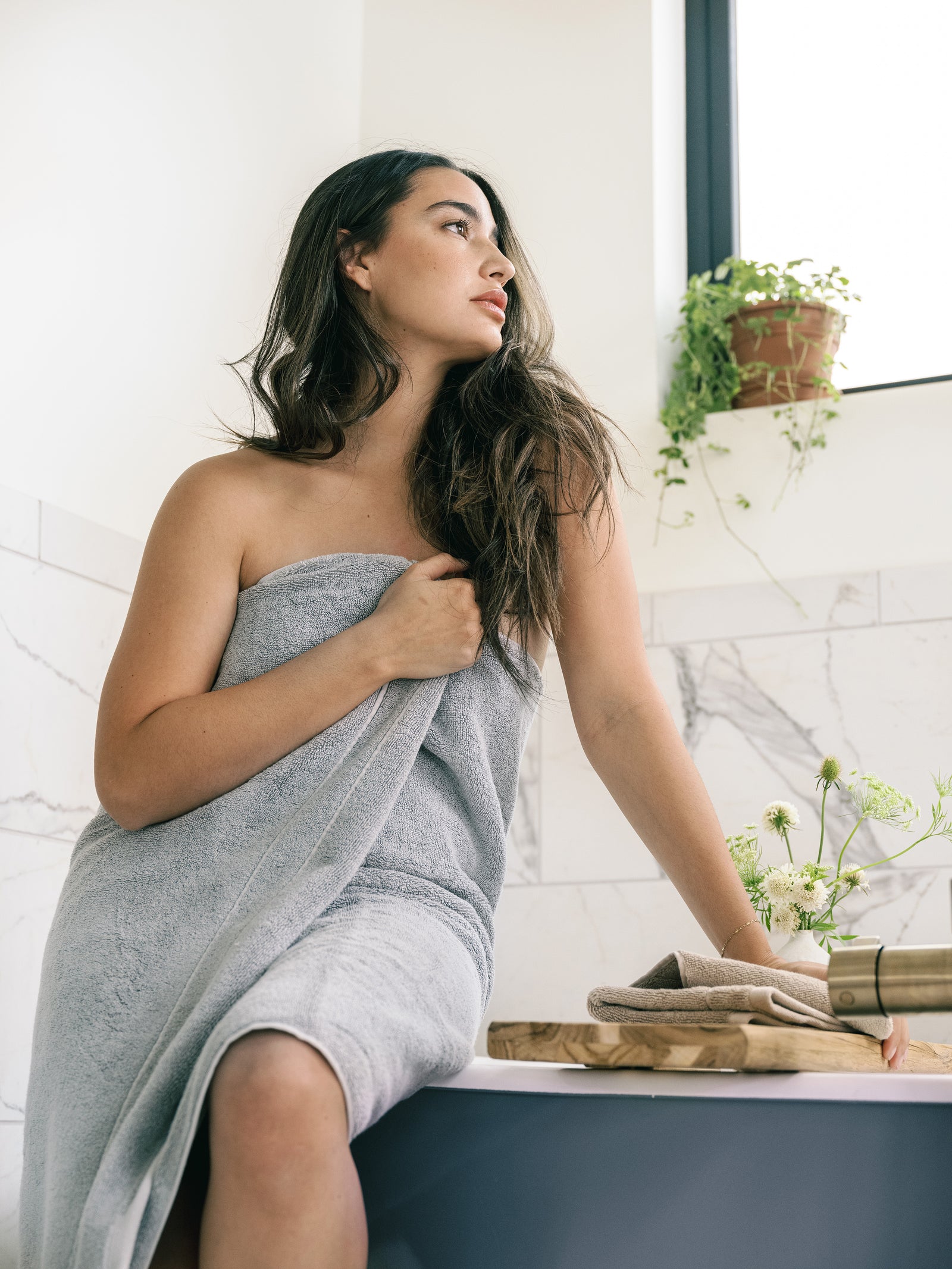Premium Plush Bath Towel in the color Harbor Mist. Photo of Harbor Mist Premium Plush Bath Towel taken in a bathroom with a white tile backsplash. There is a brunette woman holding the towel close to her body as she sits on the bathtub. 