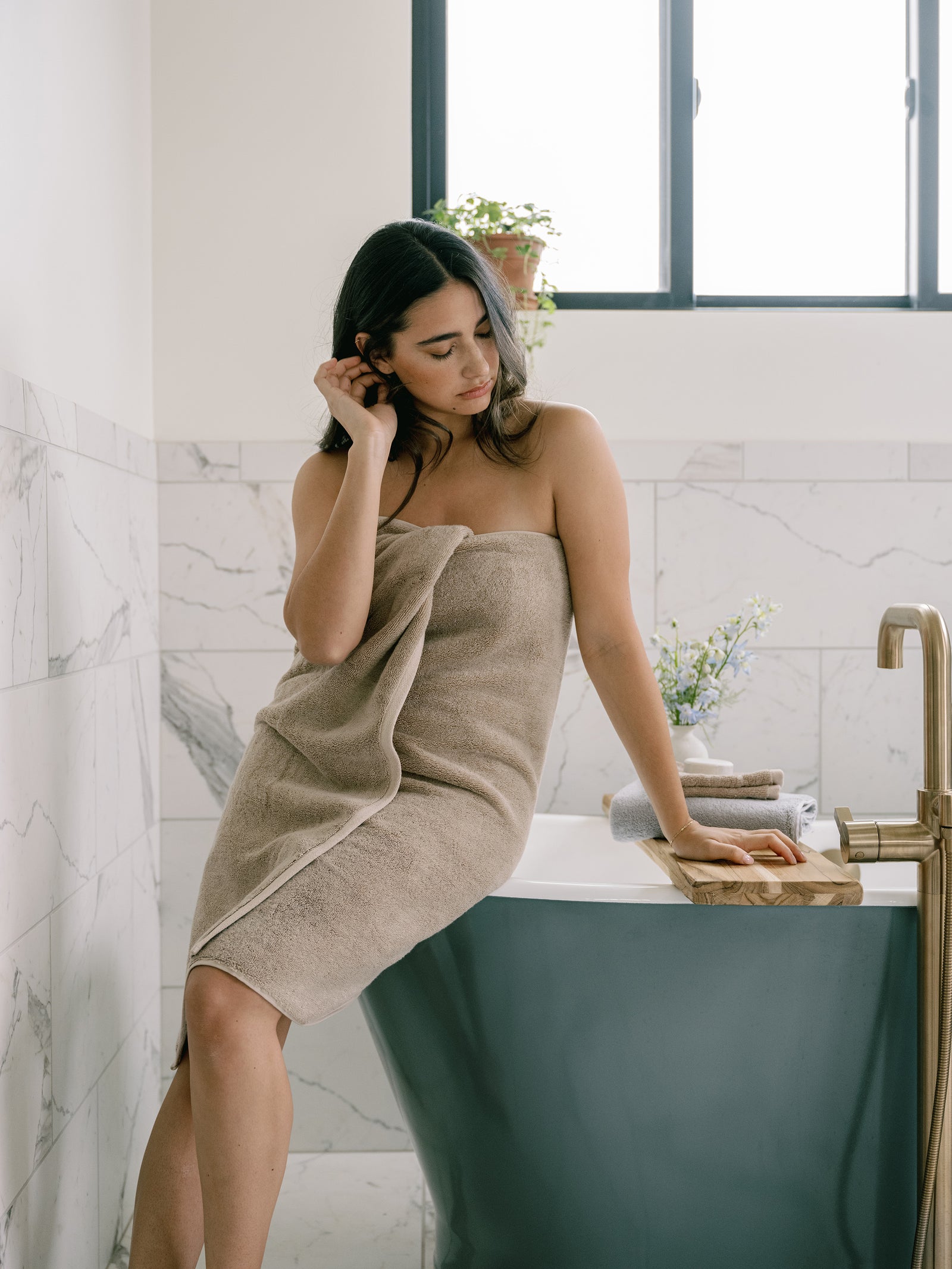 Premium Plush Bath Towel in the color Sand. Photo of Sand Premium Plush Bath Towel taken in a bathroom with a white tile backsplash. There is a brunette woman holding the towel close to her body as she sits on the bathtub. 