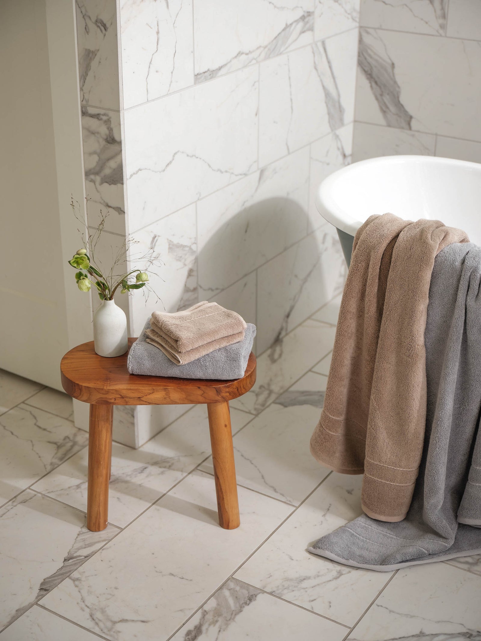 Premium Plush Hand Towels in the color Sand and Harbor Mist. Photo of Sand and Harbor Mist Premium Plush Hand Towels taken resting on a stool in a bathroom with marble tile. Sand and harbor mist premium plush bath sheets are draped over the bath tub that is in the bathroom.
