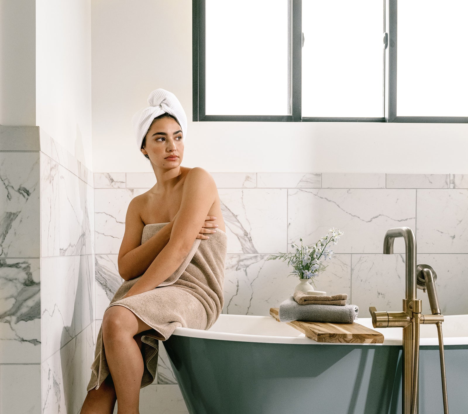 Premium Plush Bath Towel in the color Sand. Photo of Sand Premium Plush Bath Towel taken in a bathroom with a white tile backsplash. There is a brunette woman holding the towel close to her body as she sits on the bathtub. 