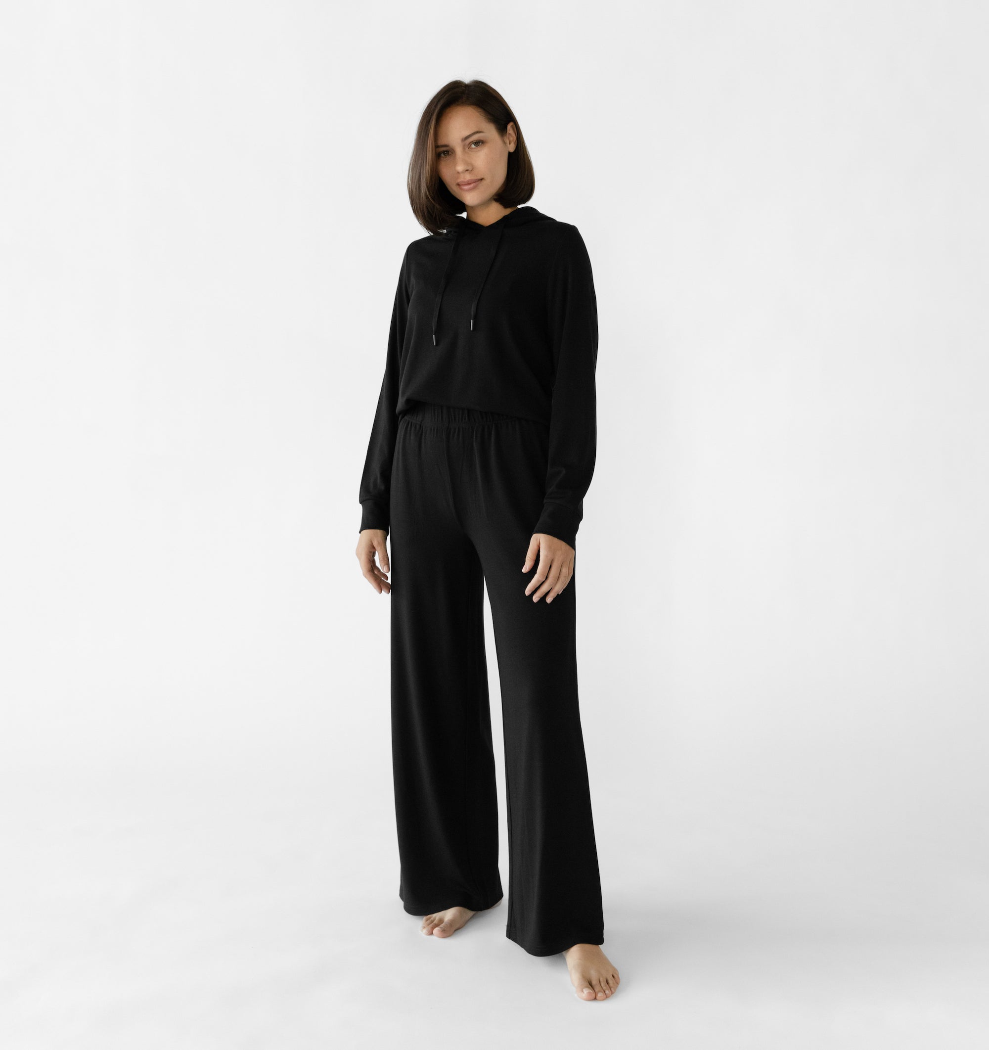 A woman stands in a relaxed pose wearing a black loungewear set from Cozy Earth, featuring the Women's Ultra-Soft Bamboo Wide Leg Pull On Pant TALL and a matching hooded sweatshirt.  