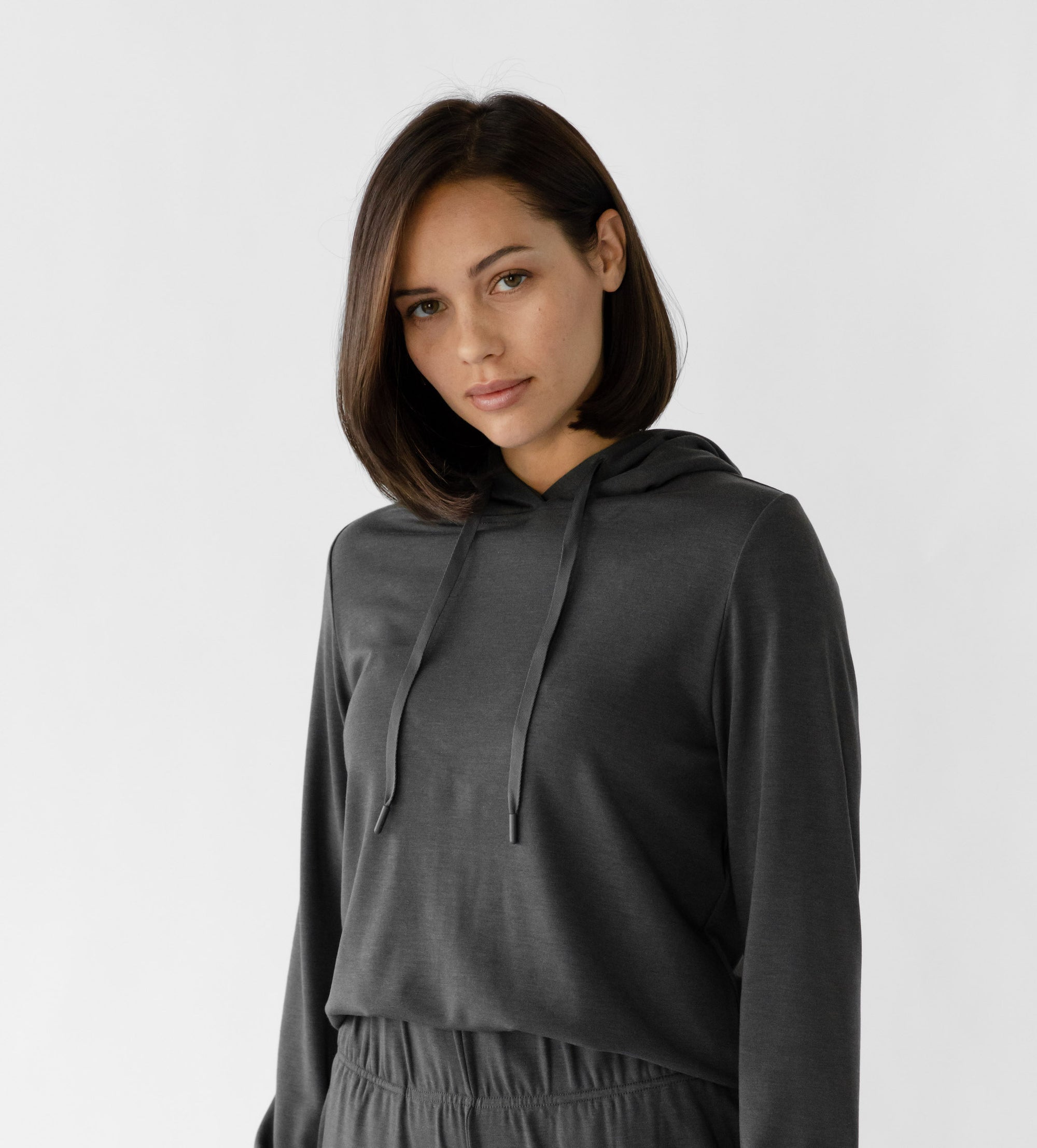 A woman wearing a grey Cozy Earth Brushed Bamboo Hoodie with a drawstring and sleeve cuffs. The hoodie has an embroidered logo on the front. 