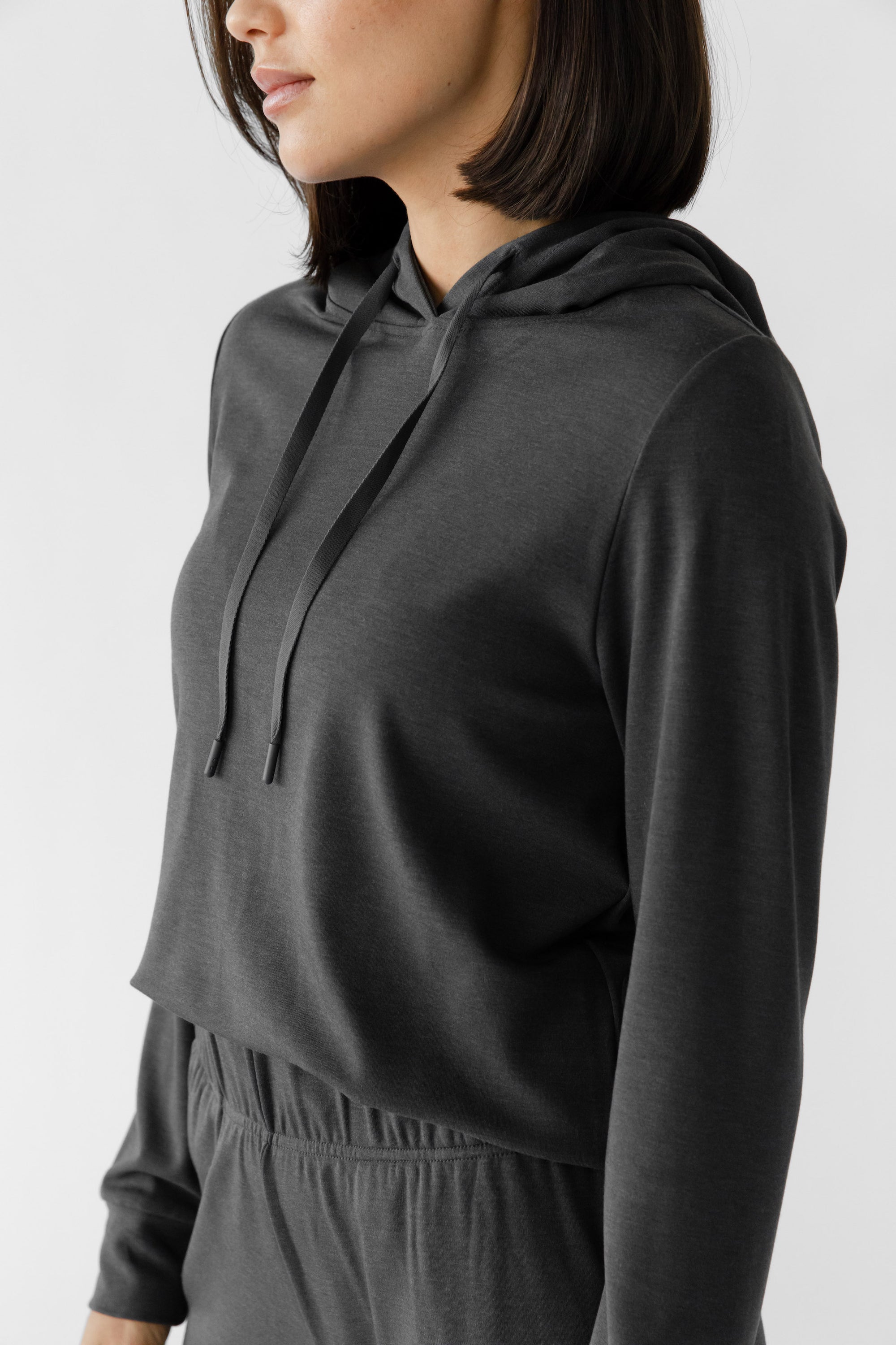 A woman wearing a grey Cozy Earth Brushed Bamboo Hoodie with a drawstring and sleeve cuffs. The hoodie has an embroidered logo on the front. 