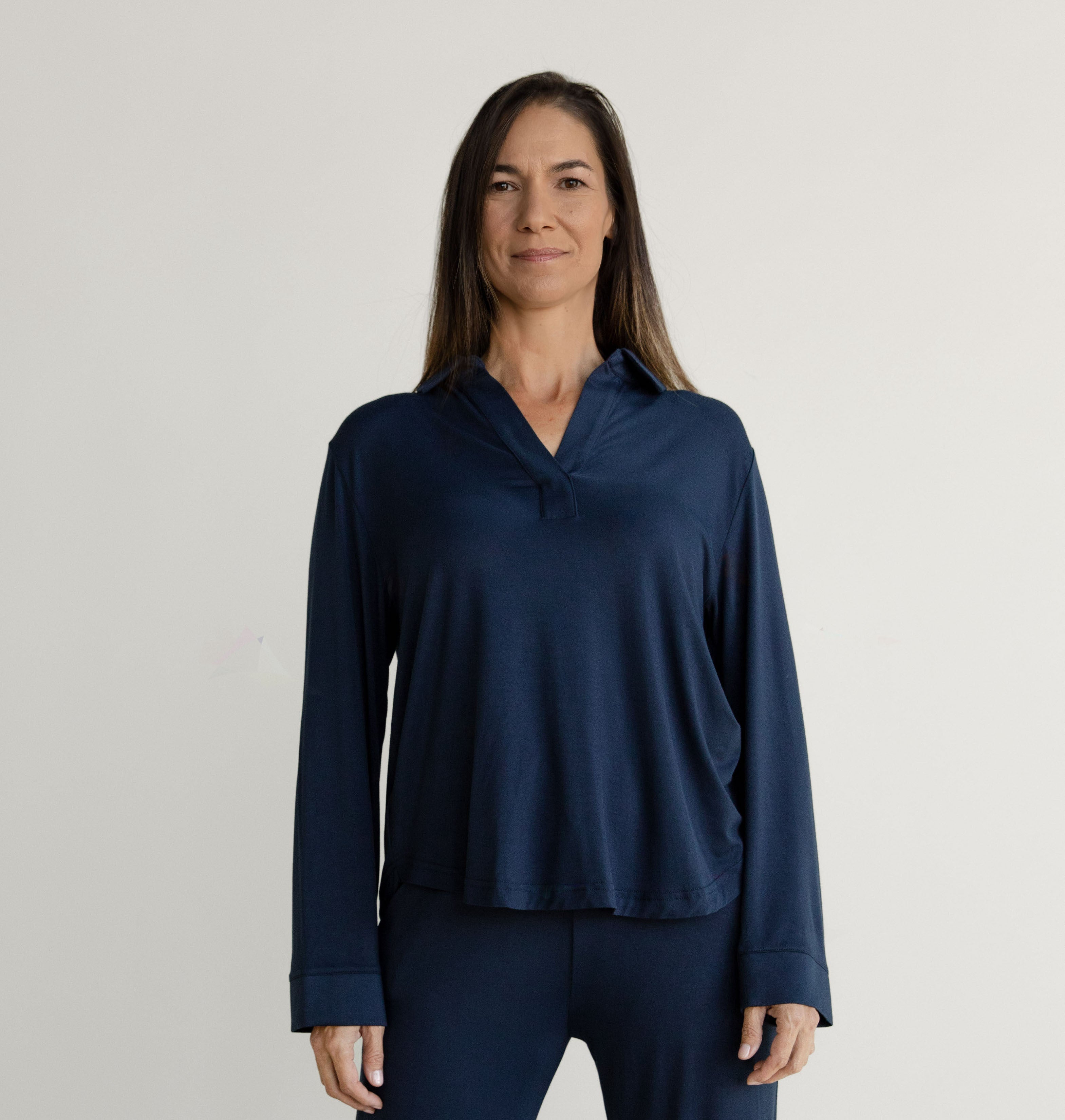 A woman with long brown hair stands against a plain, light-colored background. She is wearing the Navy Stretch Knit Bamboo Long Sleeve Polo by Cozy Earth and matching pants. She is looking forward and smiling slightly.