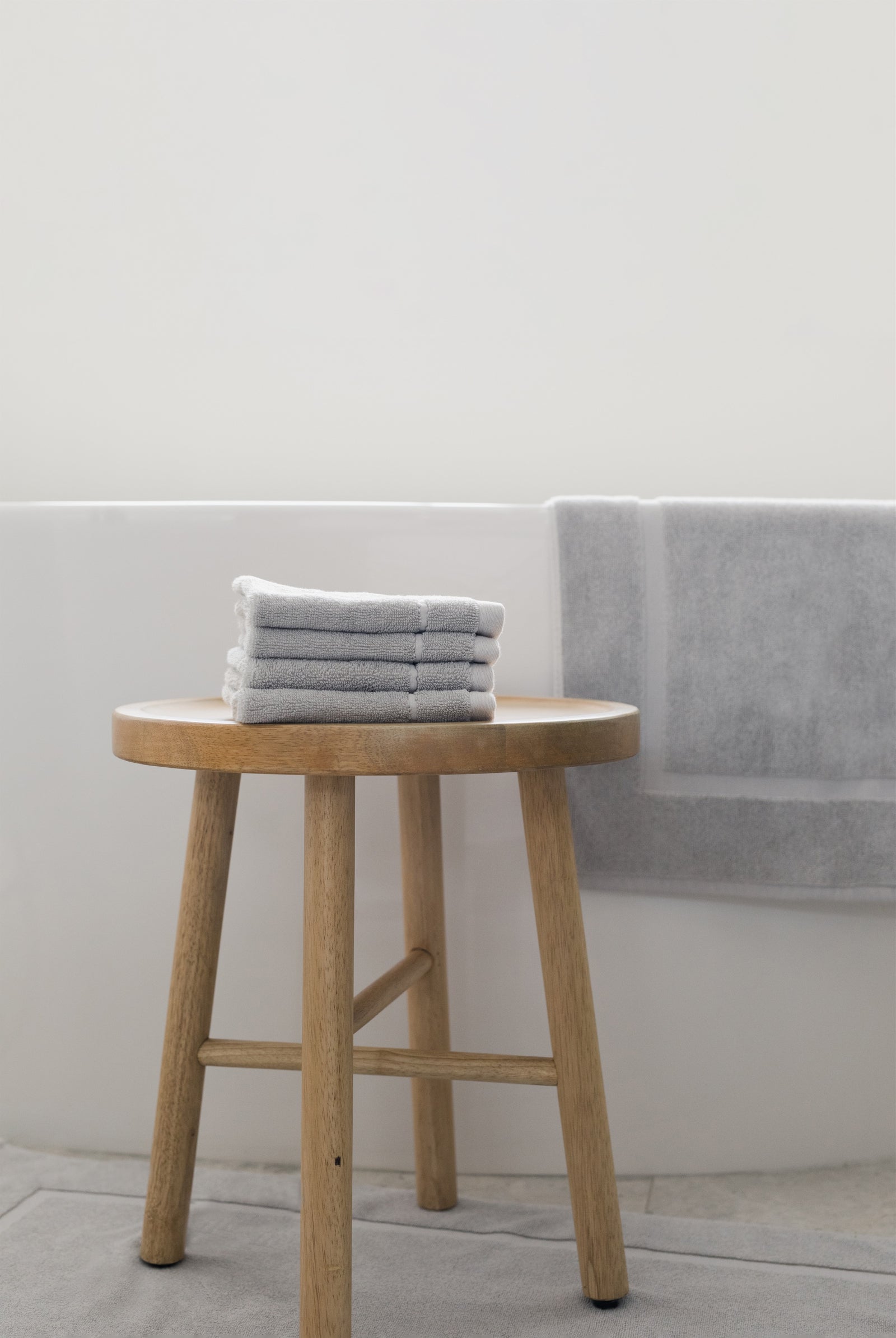 Light Grey Premium Plush Bath Mat resting on white bathtub. The photo was taken in a bathroom showing a wooden stool with wash cloths next to the bathtub.