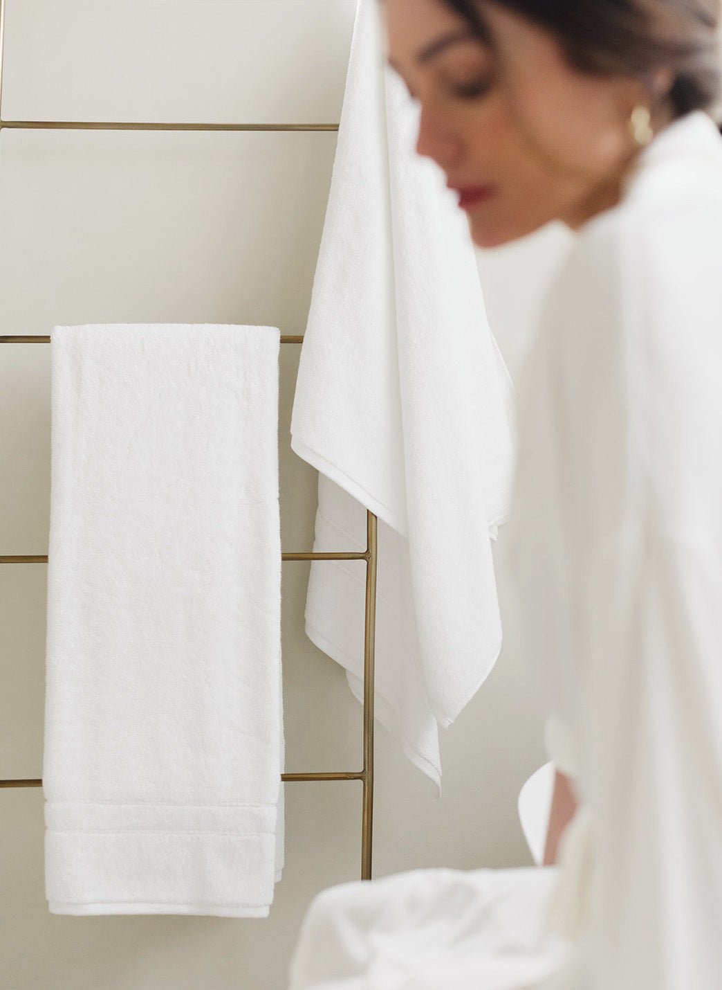Premium Plush Bath Towels in the color White. Photo of Premium Plush Bath Towels taken in a bathroom showing a woman who is out of the focus of the picture. The towels are hung from a towel rack. 