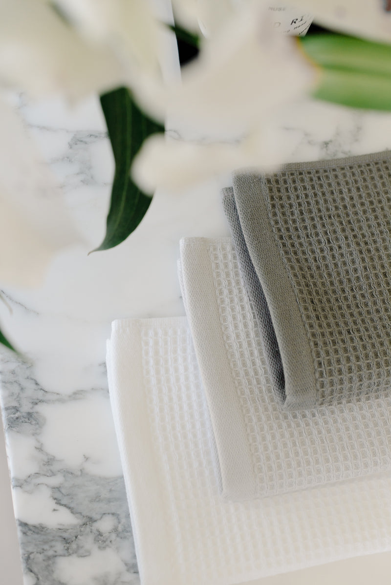 Charcoal, Light Grey, and White Waffle Wash Cloths neatly folded. The photo was taken with the cloths resting on a white marble sink.