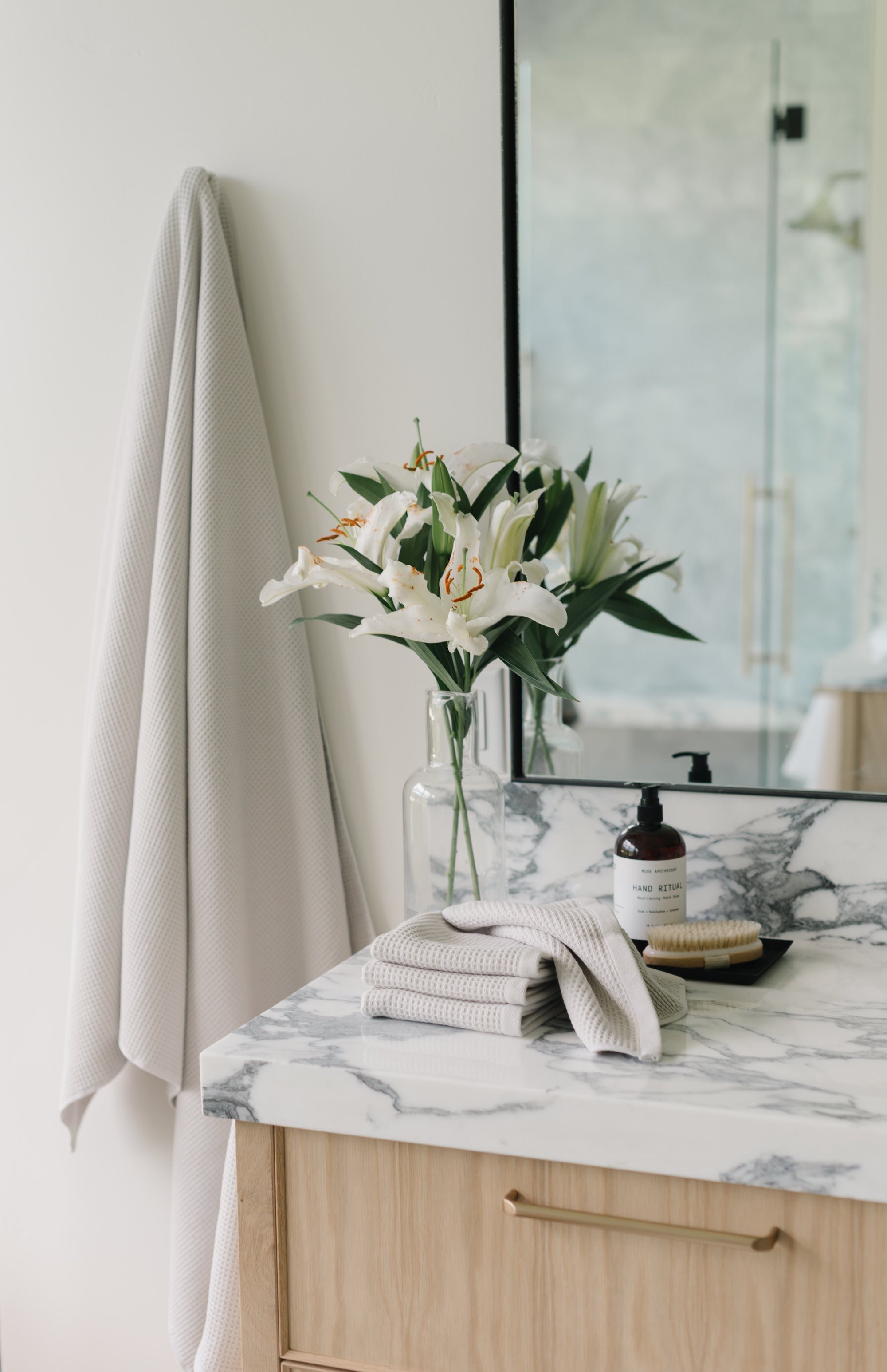 Light Grey Waffle Hand Towel hanging from a gold towel bar. The photo was taken at a white marble bathroom sink. A Waffle Bath Towel is seen hanging on the wall in the background. 