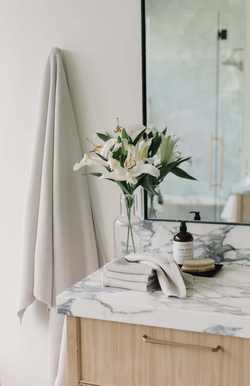 Light Grey Waffle Hand Towel hanging from a gold towel bar. The photo was taken at a white marble bathroom sink. A Waffle Bath Towel is seen hanging on the wall in the background.