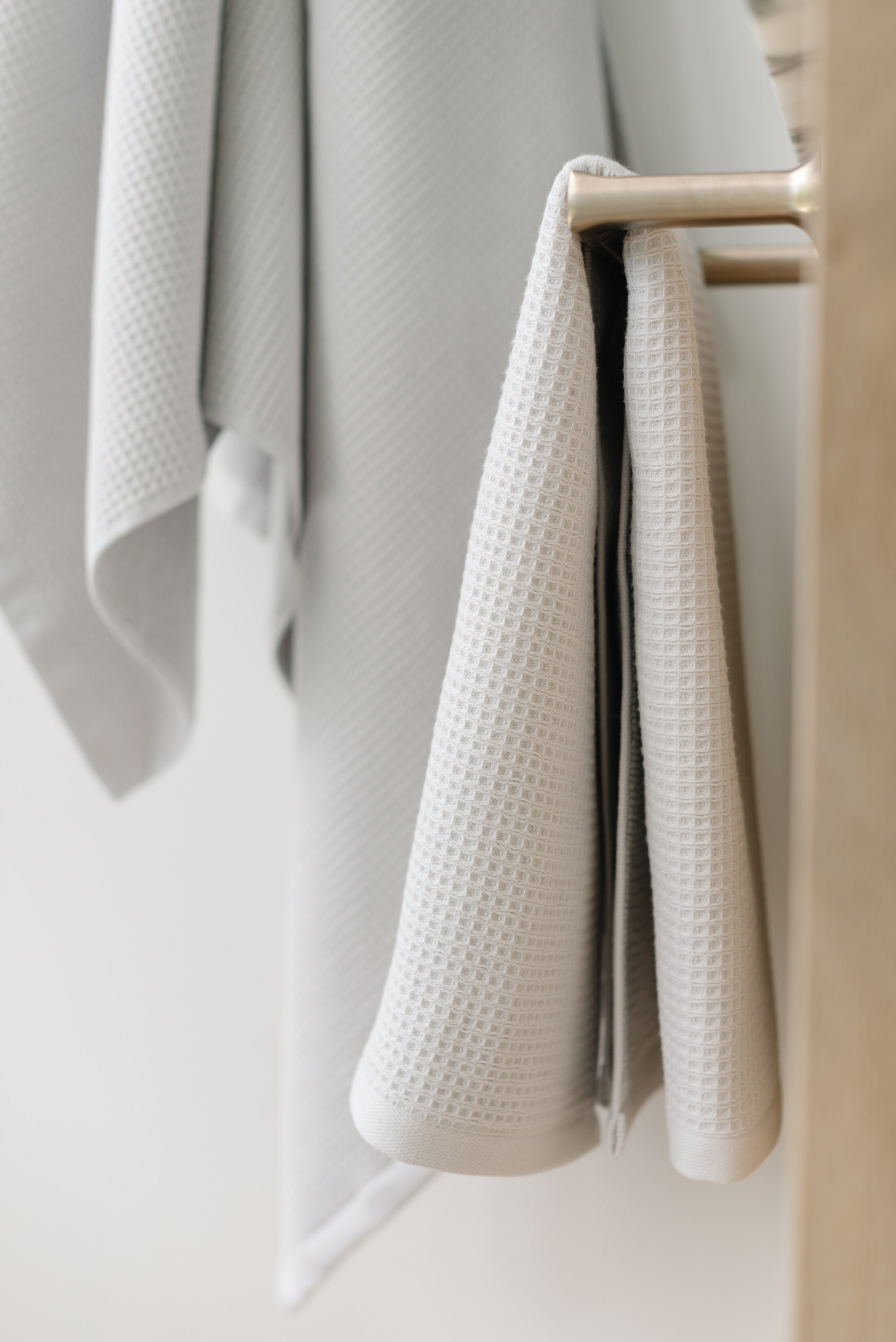 Light Grey Waffle Hang Towel handing from a gold towel bar.  The photo was taken at a white marble bathroom sink. A Waffle Bath Towel is seen hanging on the wall in the background. |Color:Light Grey